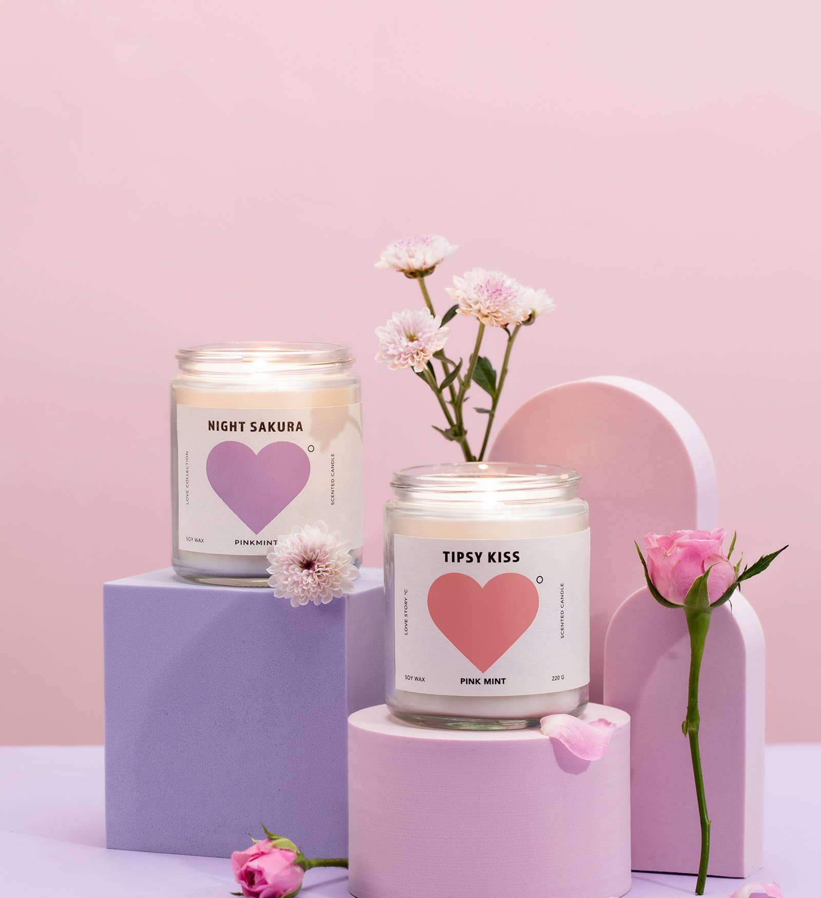 Two candles with heart designs on labels, surrounded by flowers on a pink background
