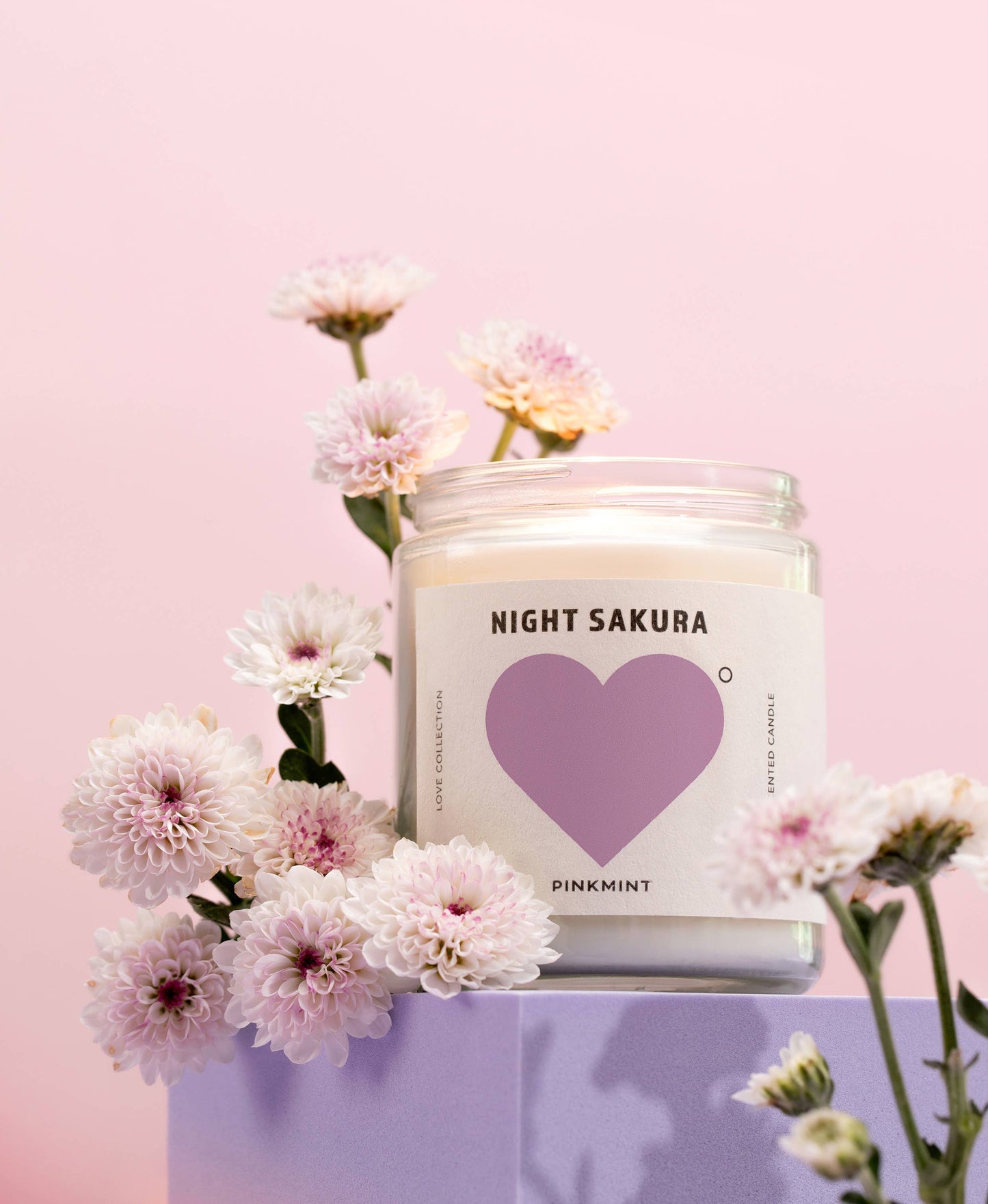 Candle labeled 'Night Sakura' with a heart design, surrounded by flowers on a pink background