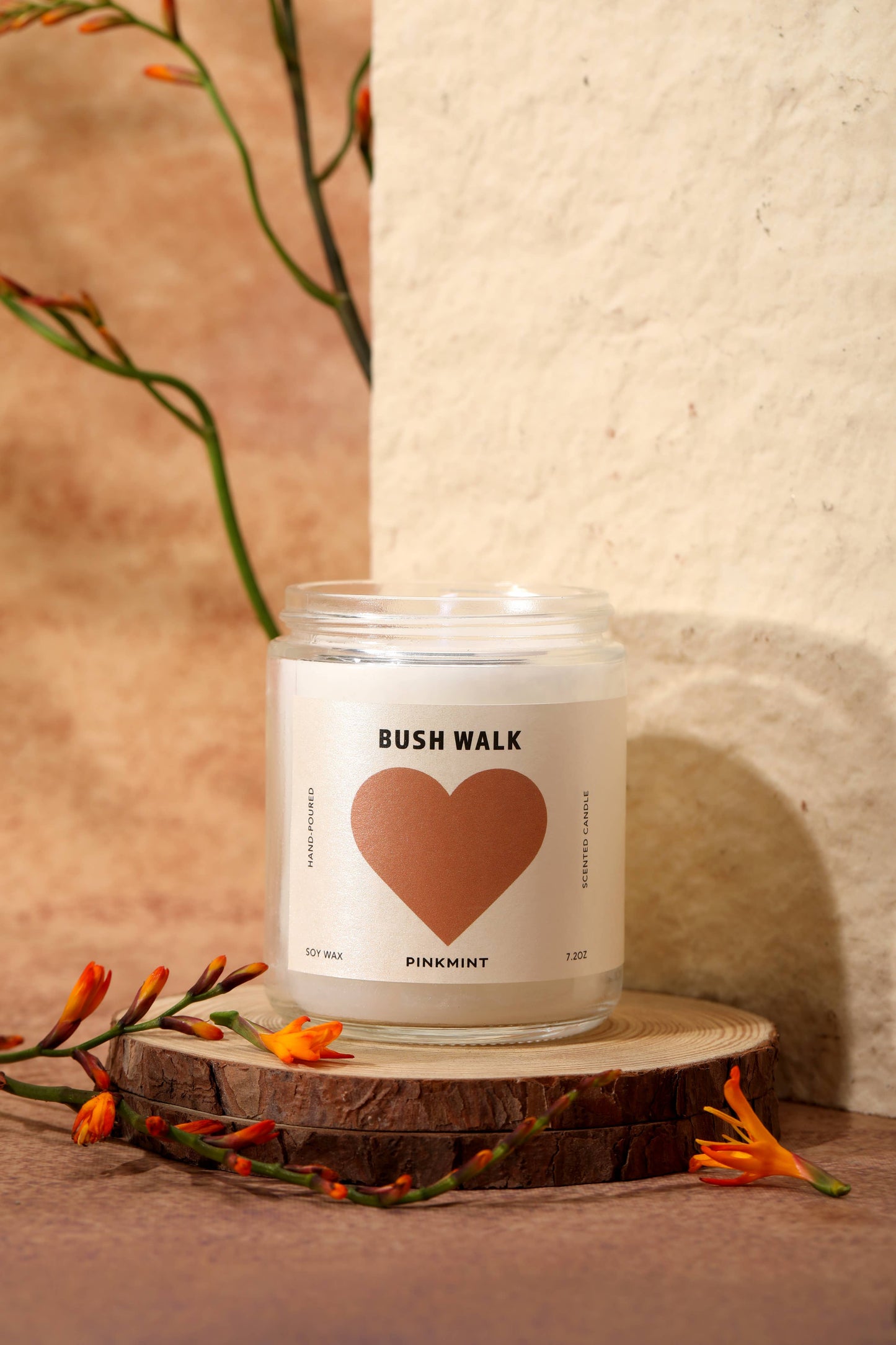 Candle labeled 'Bush Walk' with a heart design on a wooden surface with flowers.
